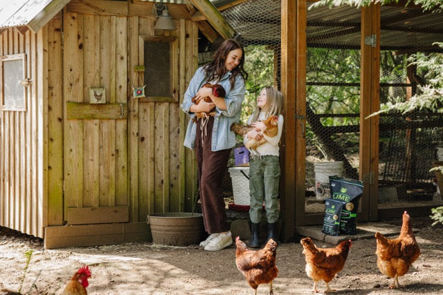 A mother and daughter both holding chickens and standing in front of a large chicken coop.