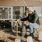 A man kneels in front of a chicken coop, using a cup to spread First Saturday Lime.