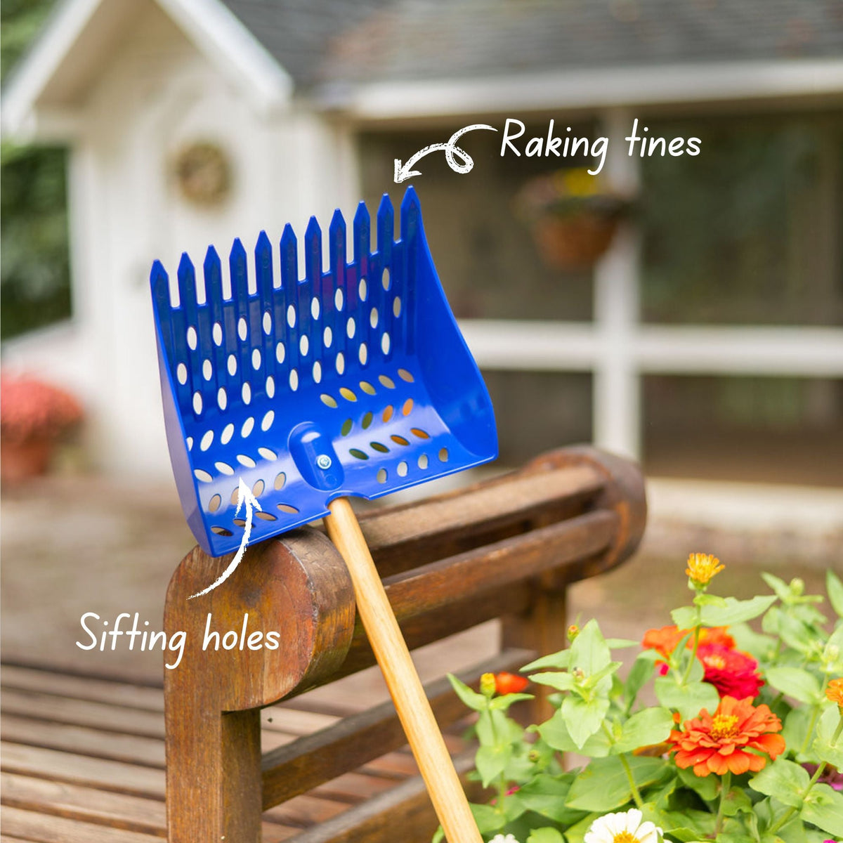A blue coop scoop with a long wooden handle resting on a bench, with flowers and a chicken coop in the background.