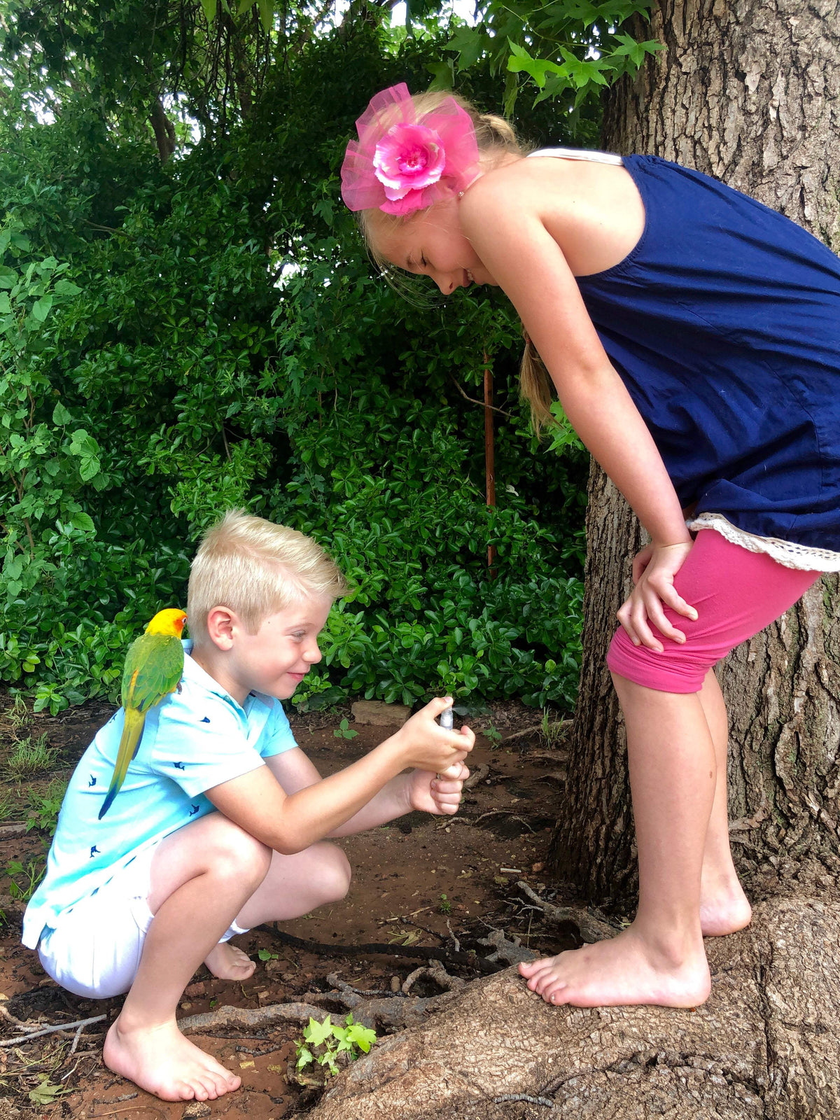 A young boy with a colorful bird on his shoulder sprays First Saturday Insect Repellent spray on a young girl.